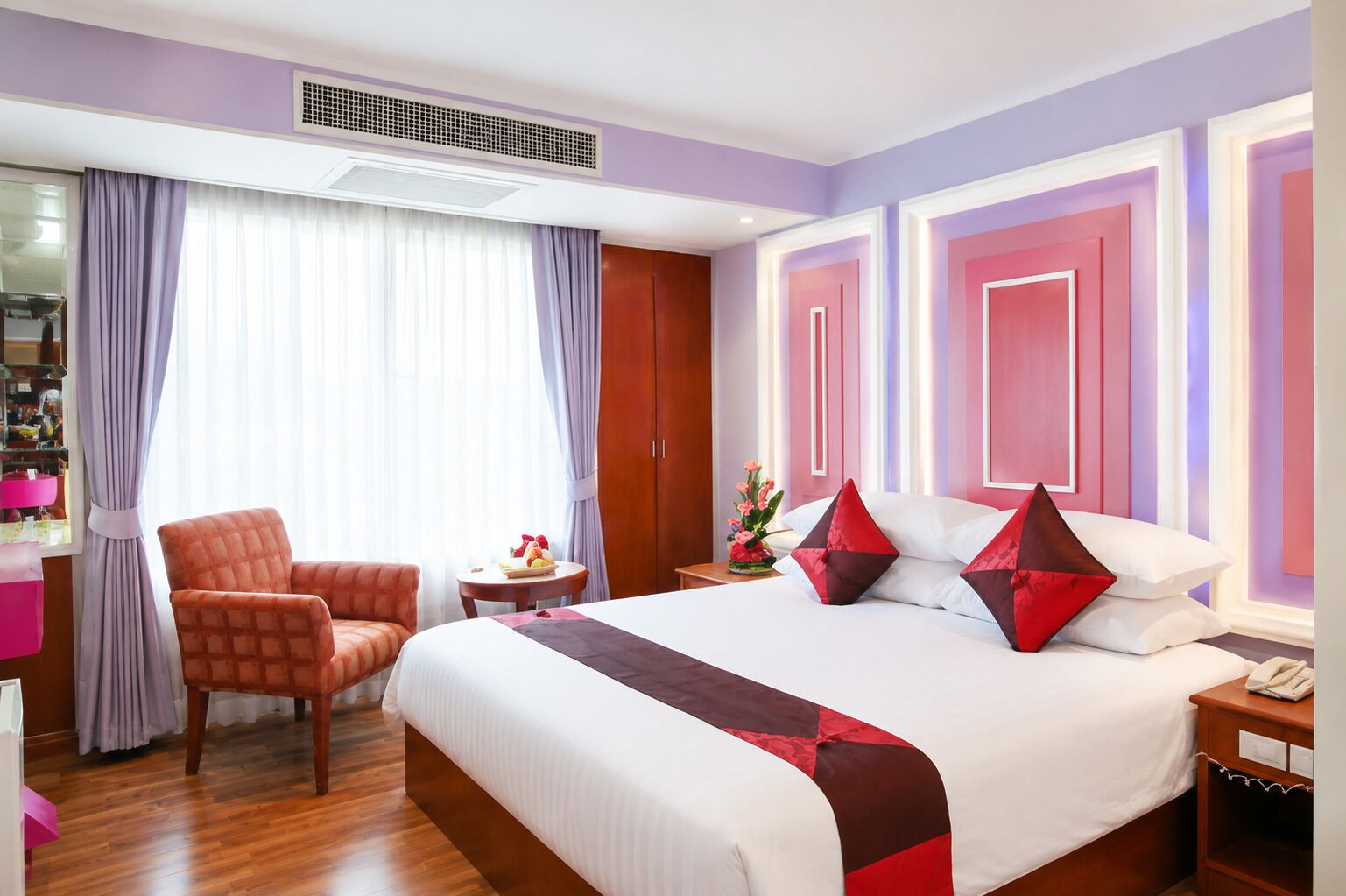 Quarto de hotel com uma cama grande, uma cadeira, uma mesa pequena com uma cesta de frutas e uma mesinha de cabeceira com um telefone.