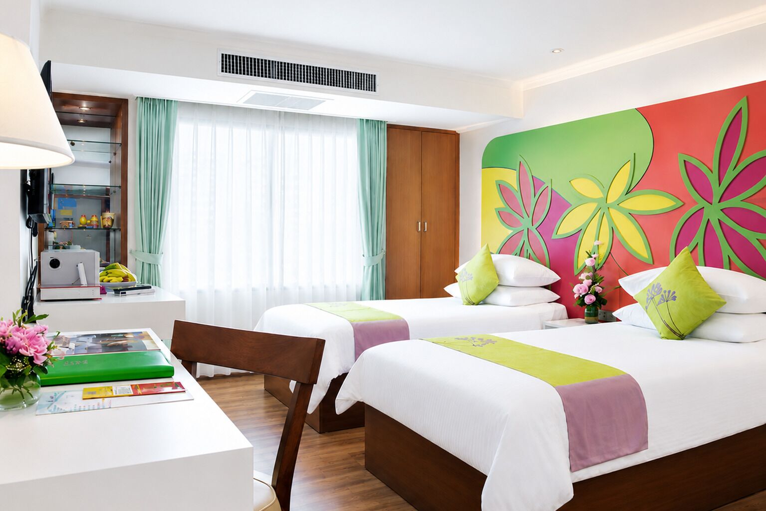 Une chambre d’hôtel avec deux lits, un bureau, une chaise, une lampe, une fenêtre avec des rideaux et une décoration murale colorée.