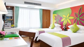 A hotel room with two beds, a desk, a chair, a lamp, a window with curtains, and a colorful wall decoration.