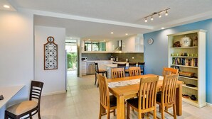 A modern kitchen with white cabinets, a dining area with wooden chairs, and a bookshelf filled with books and decorative items. - Hibiscus 006 (Whitsundays)