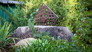 Una escultura metálica geométrica sobre una roca en un jardín exuberante.
