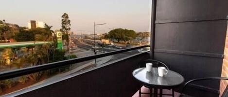Un balcón con mesa y sillas, con vista a una calle concurrida, edificios y vegetación.