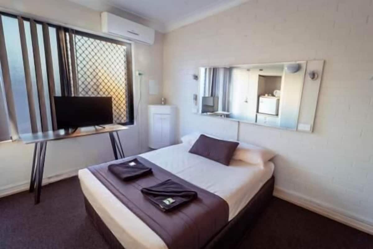 A hotel room with a bed, a TV on a desk, a mirror on the wall, and a window with blinds.