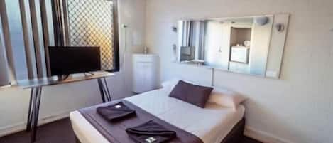 A hotel room with a bed, a TV on a desk, a mirror on the wall, and a window with blinds.