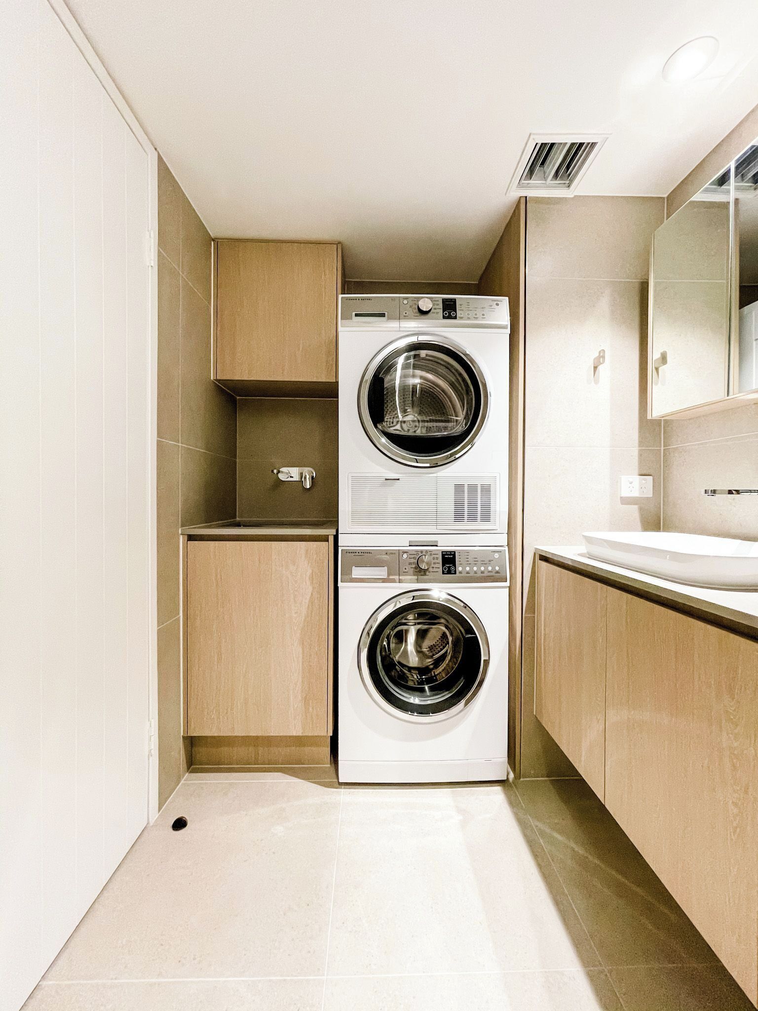 A compact laundry room with two stacked front-loading washing machines, a small cabinet, and a sink with a mirror above it.
