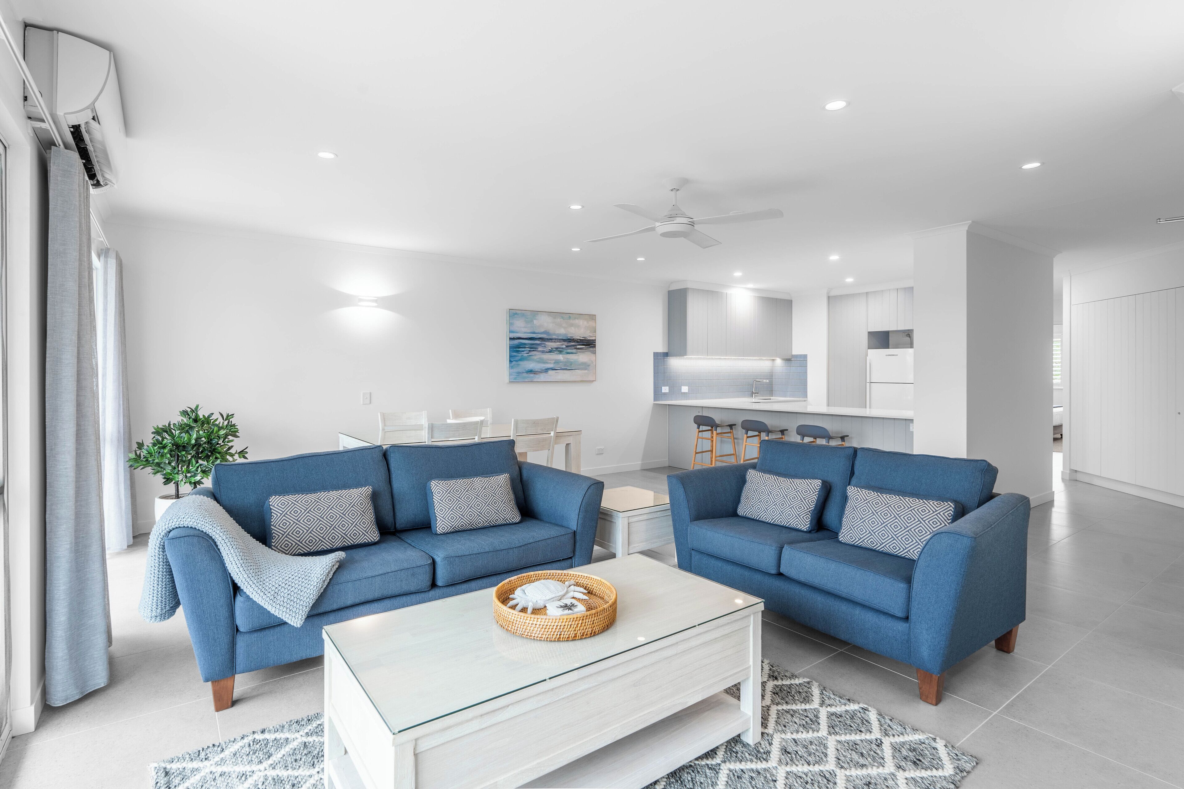 A modern living room with two blue sofas, a white coffee table, and a kitchen area in the background.