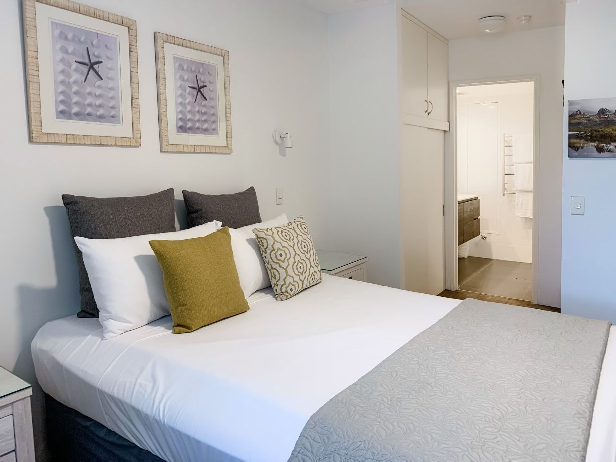 A neatly made bed with a white comforter and several pillows, a framed picture on the wall, and a visible bathroom door.
