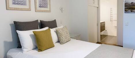A neatly made bed with a white comforter and several pillows, a framed picture on the wall, and a visible bathroom door.
