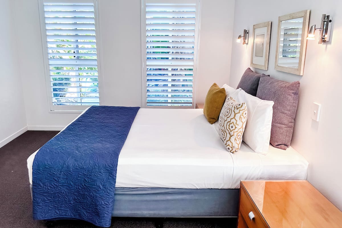 A hotel room with a bed, two windows with blinds, a bedside table, and wall-mounted lights.