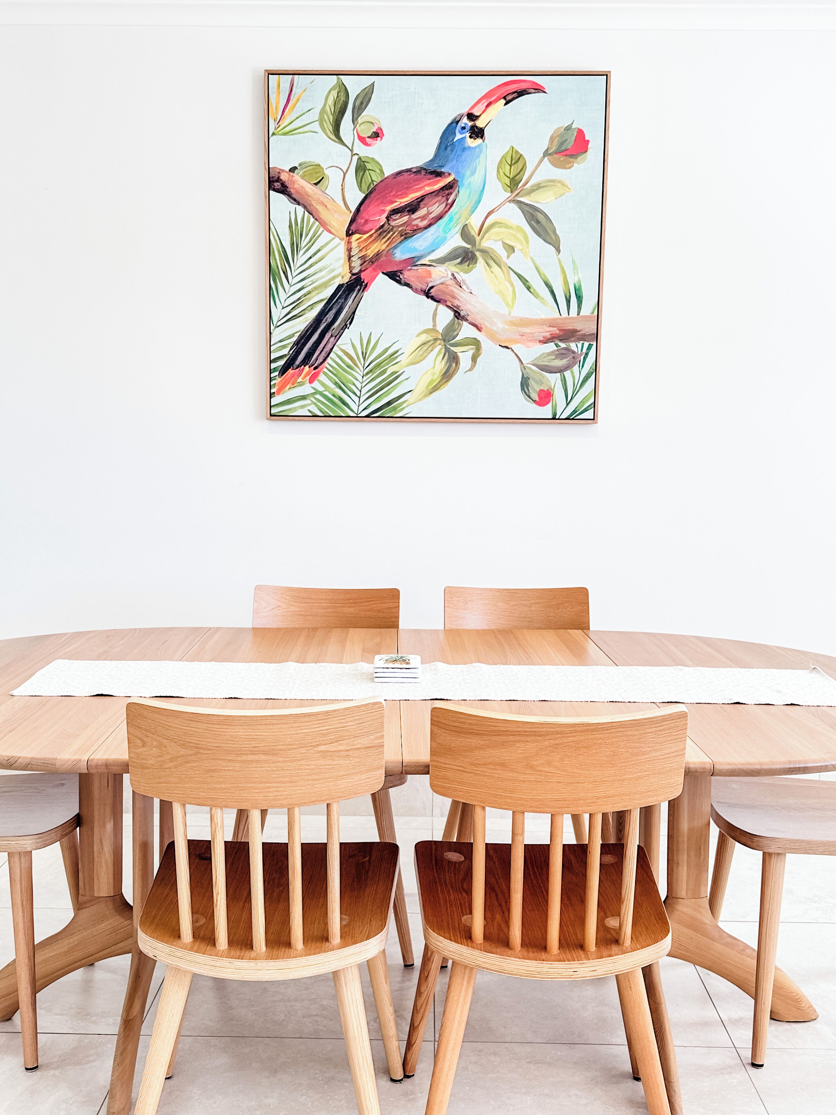 A dining table with six wooden chairs, a framed tropical bird painting on the wall, and a white placemat on the table.