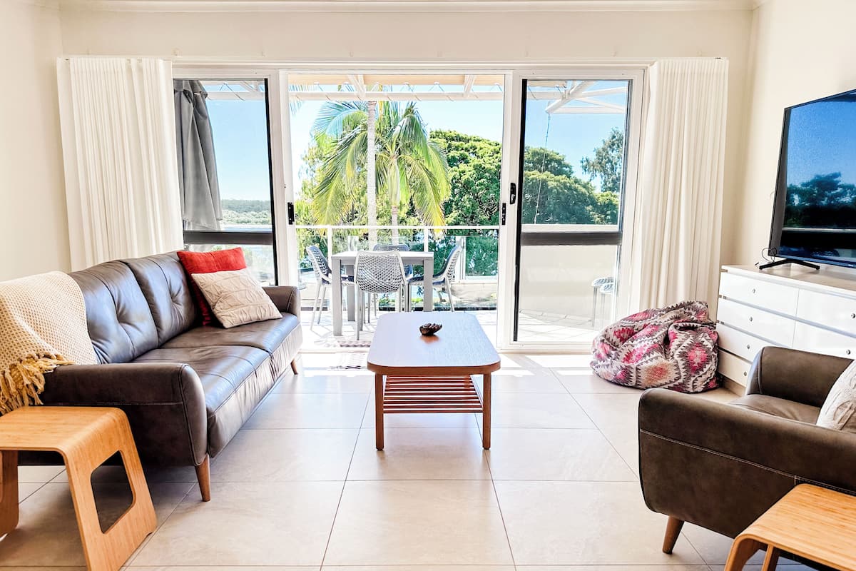 A living room with a sofa, a coffee table, and a TV. There is a balcony with a view of palm trees.