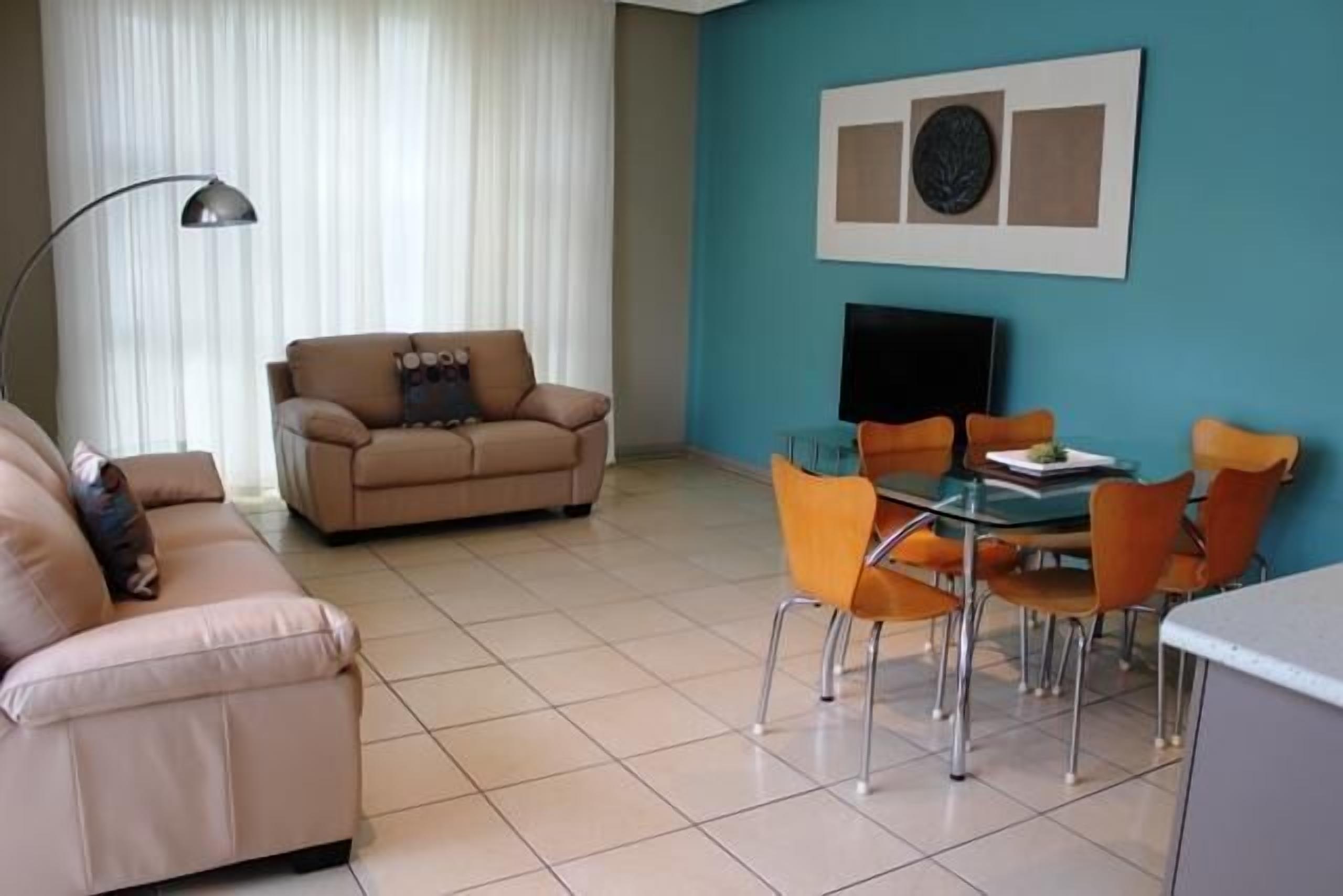 Un salón con sofá color beige, una mesa de comedor de vidrio con sillas naranjas, un televisor de pantalla plana y una pared azul decorada con obras de arte.