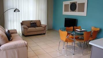 Un salón con sofá color beige, una mesa de comedor de vidrio con sillas naranjas, un televisor de pantalla plana y una pared azul decorada con obras de arte.