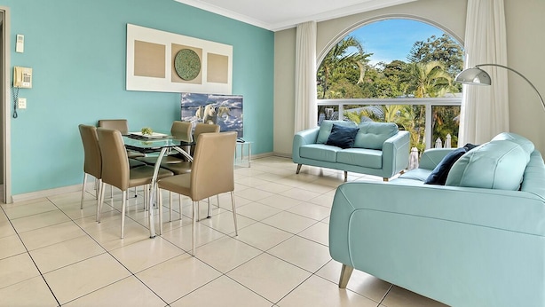 A modern living room with a dining area, a light blue sofa, a glass table, and a large window with a view of greenery. - Korora Bay Village Resort (Korora)