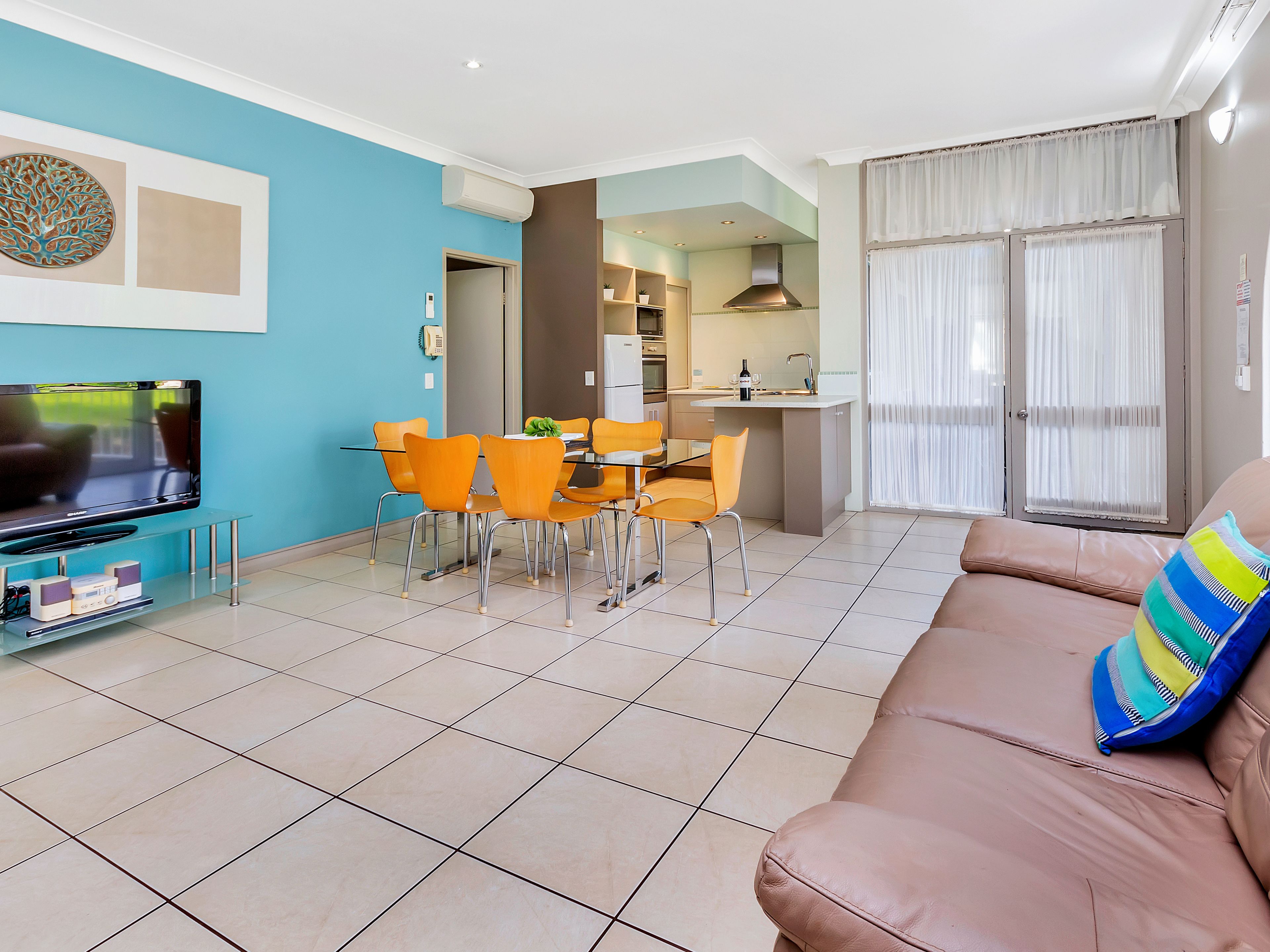 A modern living room with a TV, orange chairs, a dining table, and a sofa with a colorful pillow.