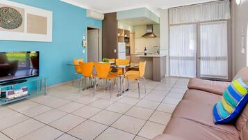 A modern living room with a TV, orange chairs, a dining table, and a sofa with a colorful pillow.