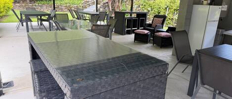 Un espacio de estar al aire libre con techo, una mesa larga y sillas, rodeado de follaje y un muro de piedra.