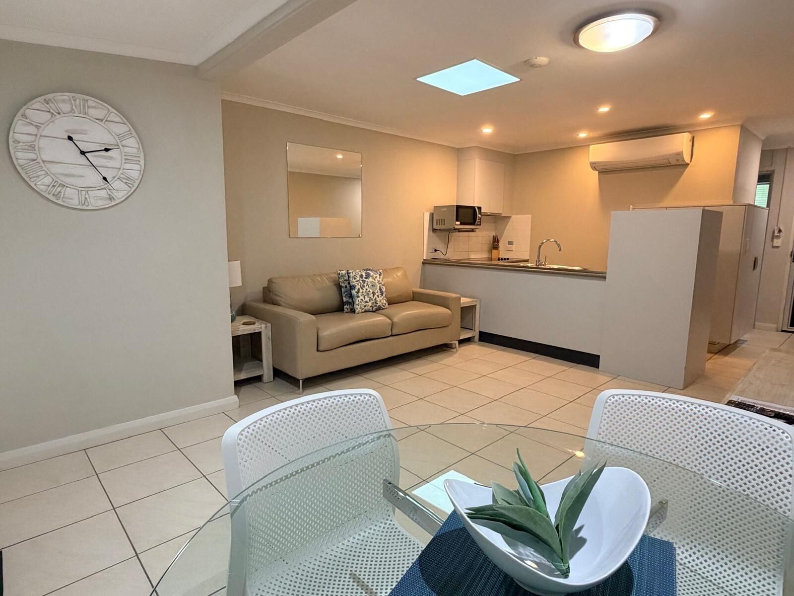 Un salon moderne avec un canapé, une table à manger et des chaises, un coin cuisine équipé d’un four à micro-ondes, et une horloge au mur.