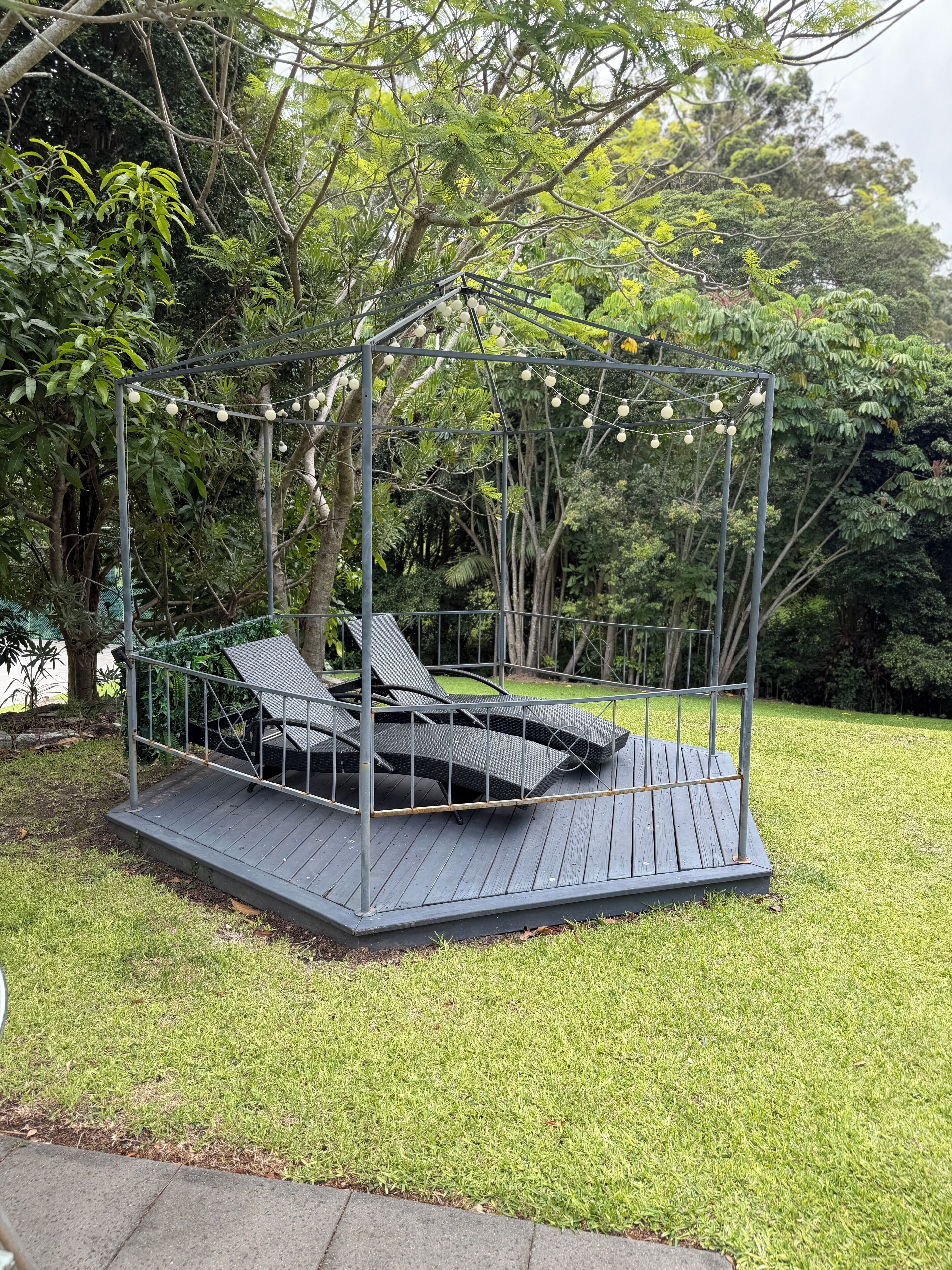 A garden swing set with a metal frame and wooden platform, surrounded by lush greenery and trees.