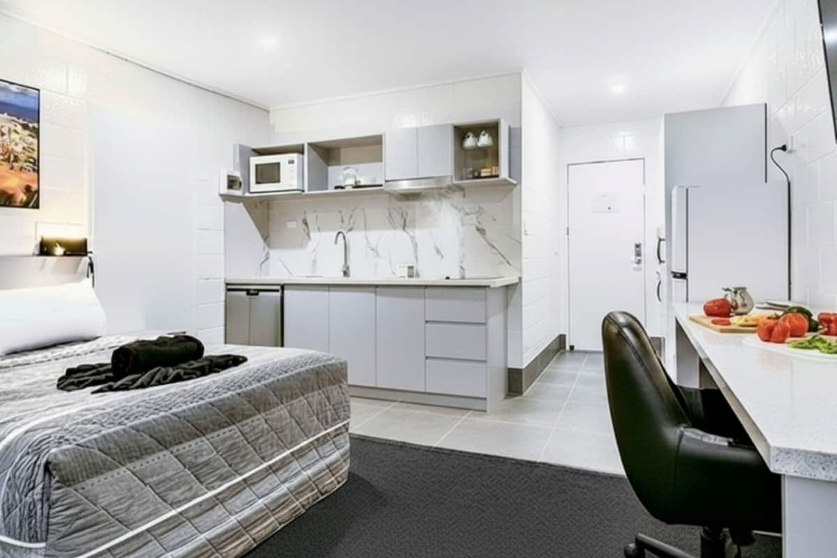 A compact hotel room with a bed, a small kitchenette, a desk with a chair, and a bathroom.