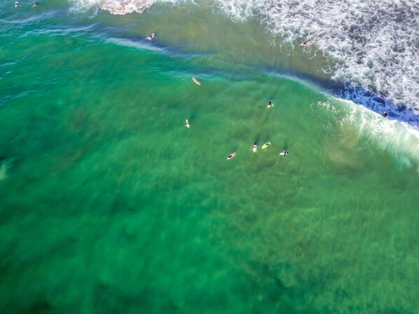 An aerial view of a beach with clear turquoise water, people swimming and surfing, and waves breaking near the shore.