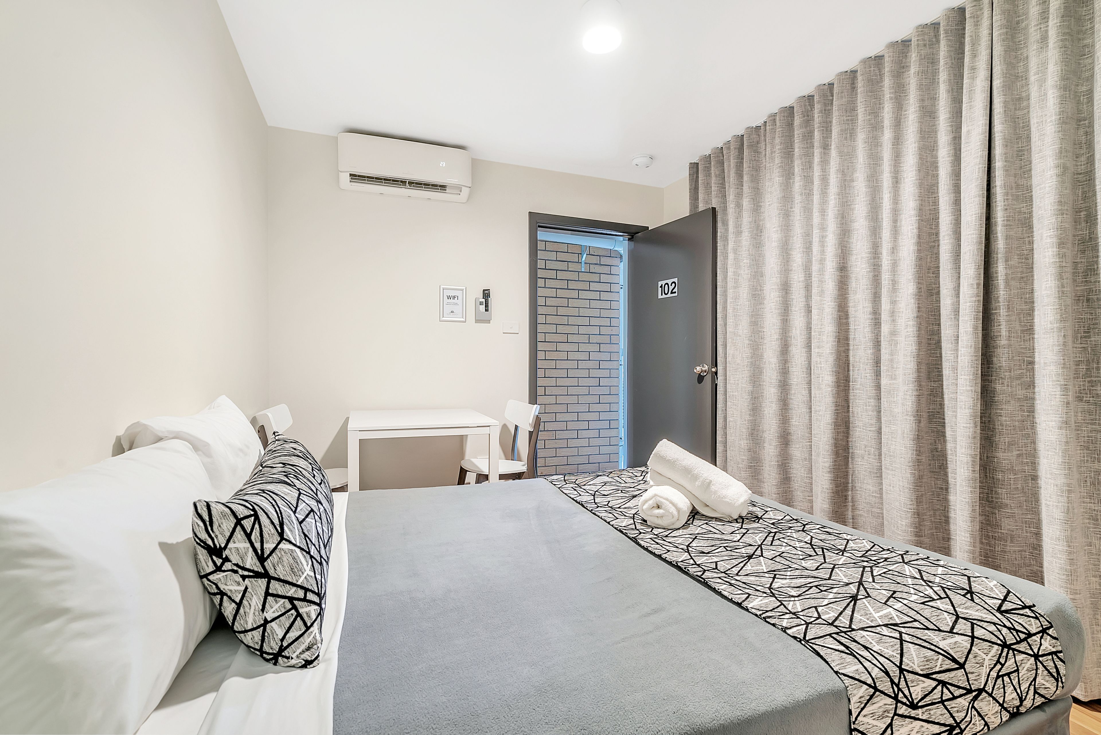 A hotel room with a bed, a desk, and a chair. There is a door with the number 102, an air conditioning unit, and a window with curtains.
