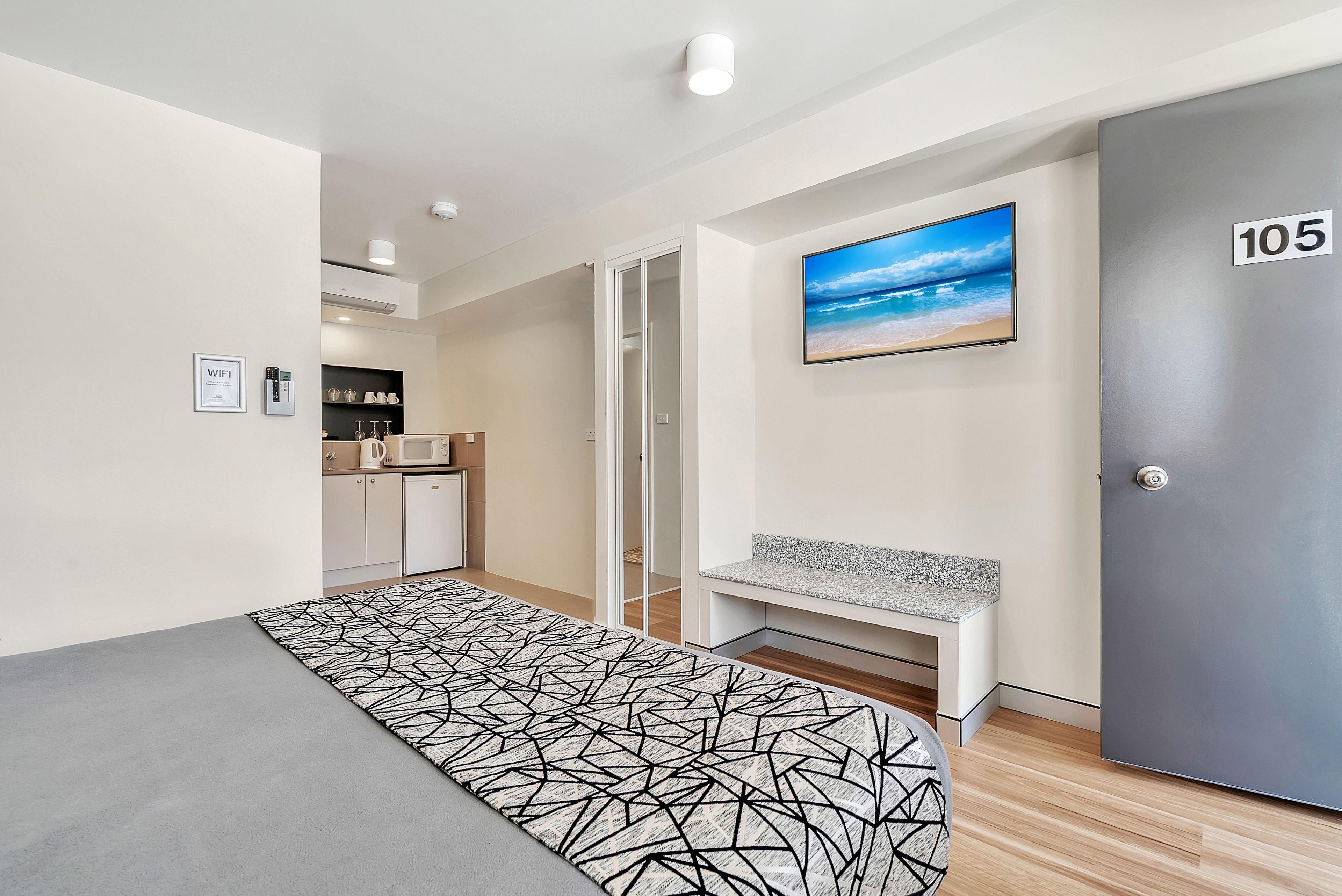 A modern hotel room with a bed, a bench, a TV, and a door marked 105.
