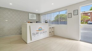 A reception desk with a logo, a window with blinds, and a view of the outside. - Surf Beach Motel Port (Port Macquarie)