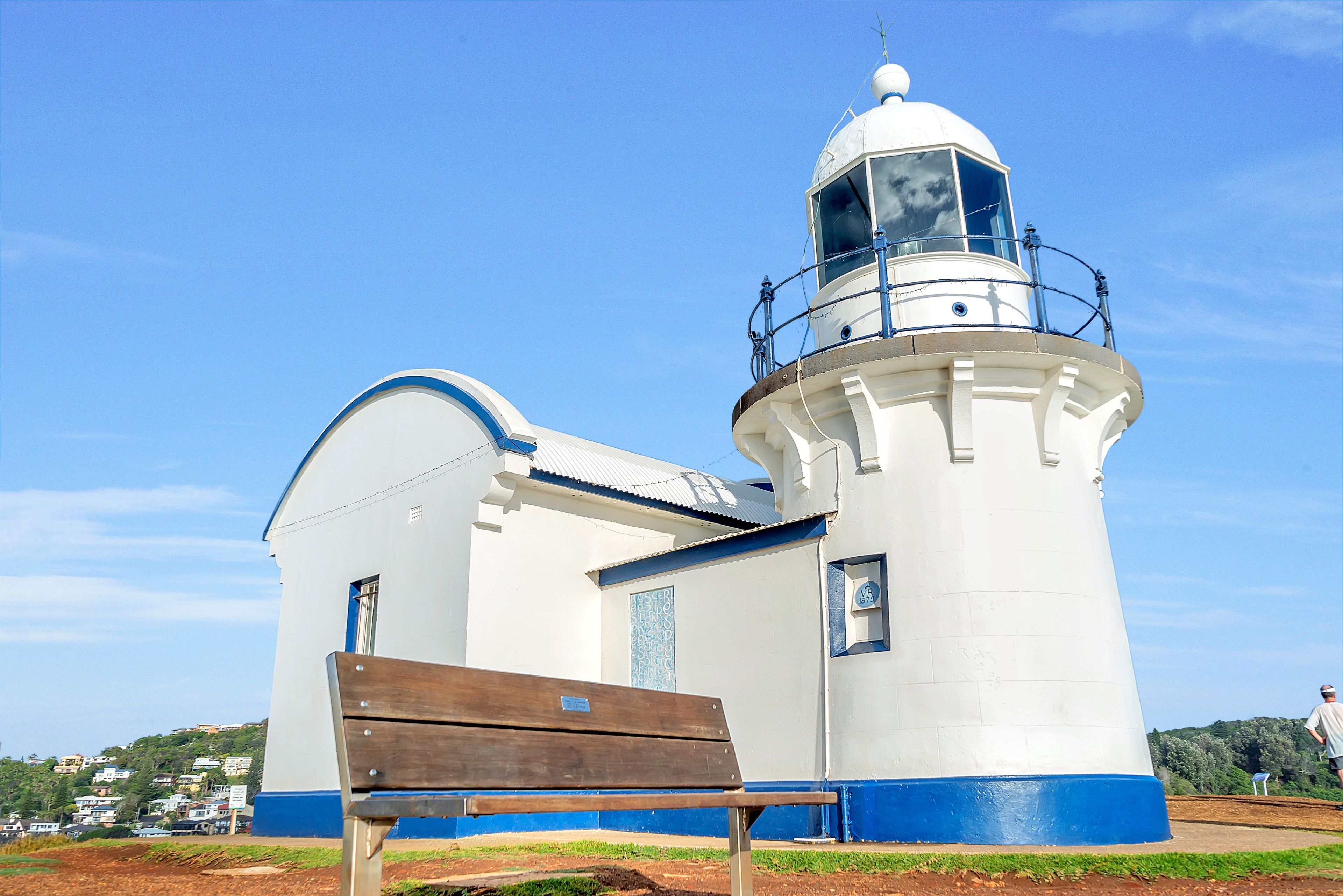 A lighthouse with a white and blue exterior, a wooden bench in front, and a clear blue sky in the background.