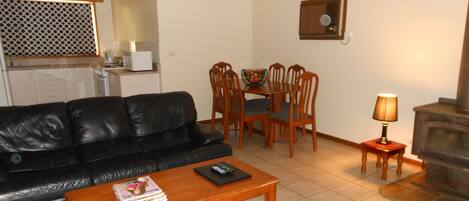 A living room with a black leather sofa, a wooden coffee table with magazines, a dining table with chairs, a small side table with a lamp, and a kitchen area in the background.