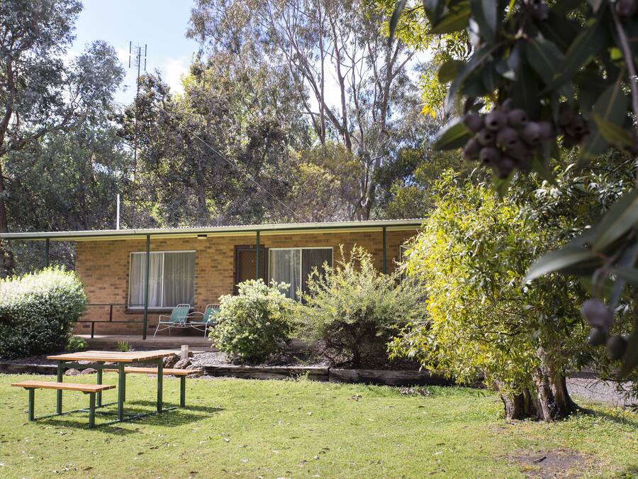 A small, single-story building with a brick exterior and a flat roof. There is a patio area with a wooden table and benches, surrounded by various shrubs and trees.