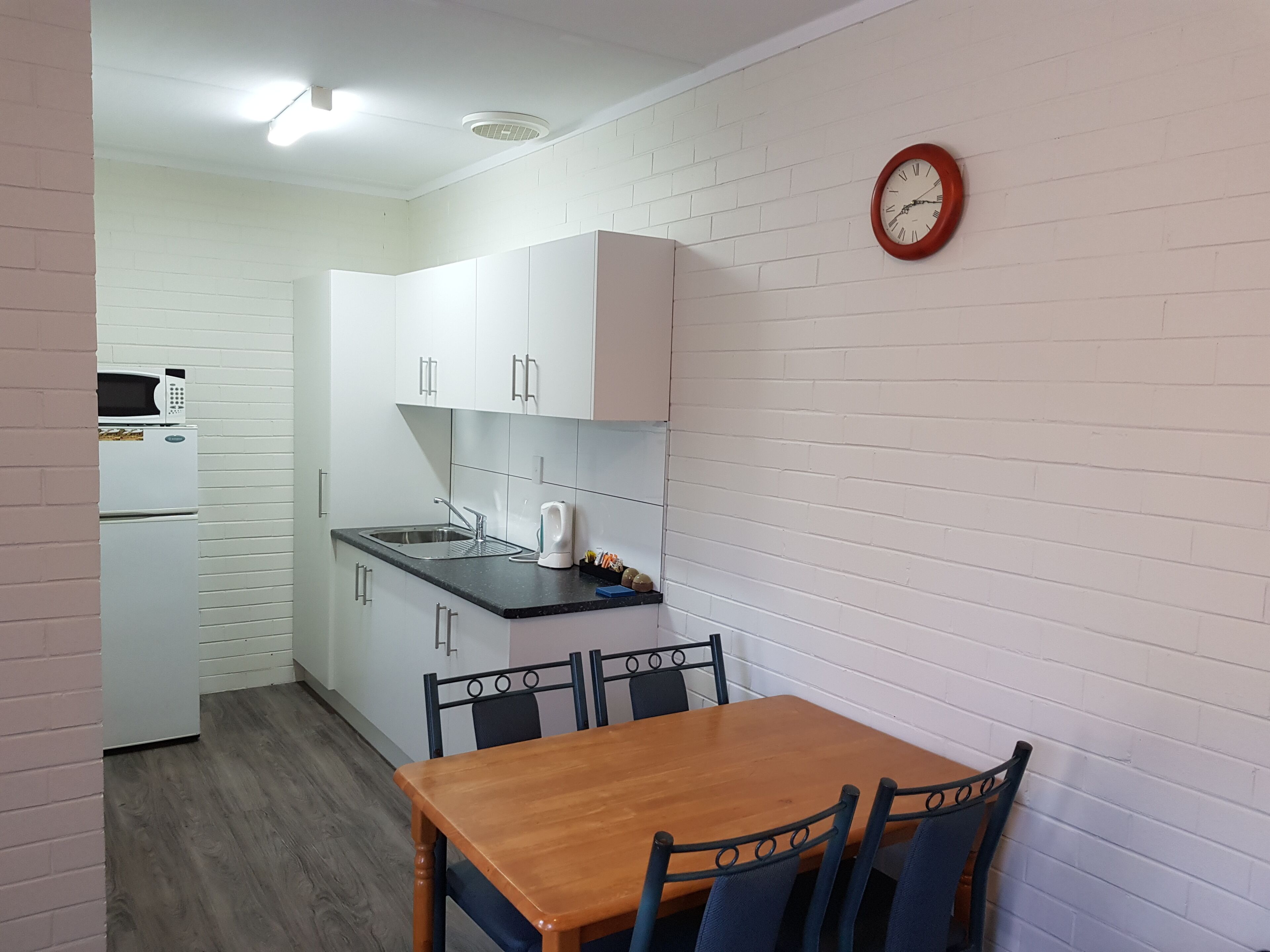 Cocina compacta con armarios blancos, microondas, tetera y fregadero. Hay una mesa de comedor de madera con sillas.