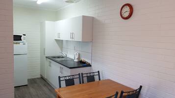 A compact kitchen with white cabinets, a microwave, a kettle, and a sink. There is a wooden dining table with chairs.