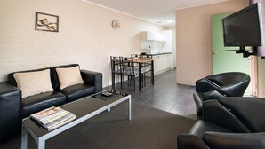 Uma sala de estar moderna com um sofá de couro preto, uma mesa de centro com revistas, uma área de jantar com mesa e cadeiras, e uma cozinha ao fundo.
