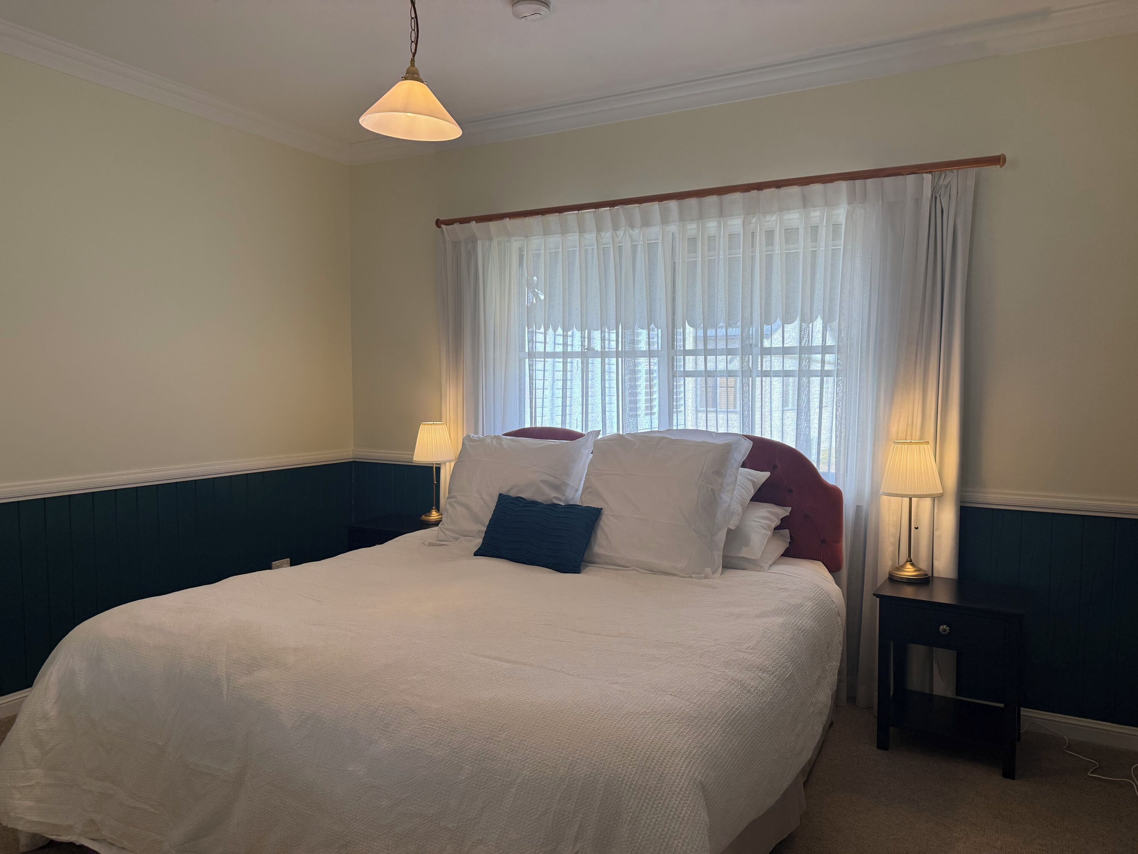 A bedroom with a bed, two bedside lamps, a window with curtains, and a hanging light fixture.