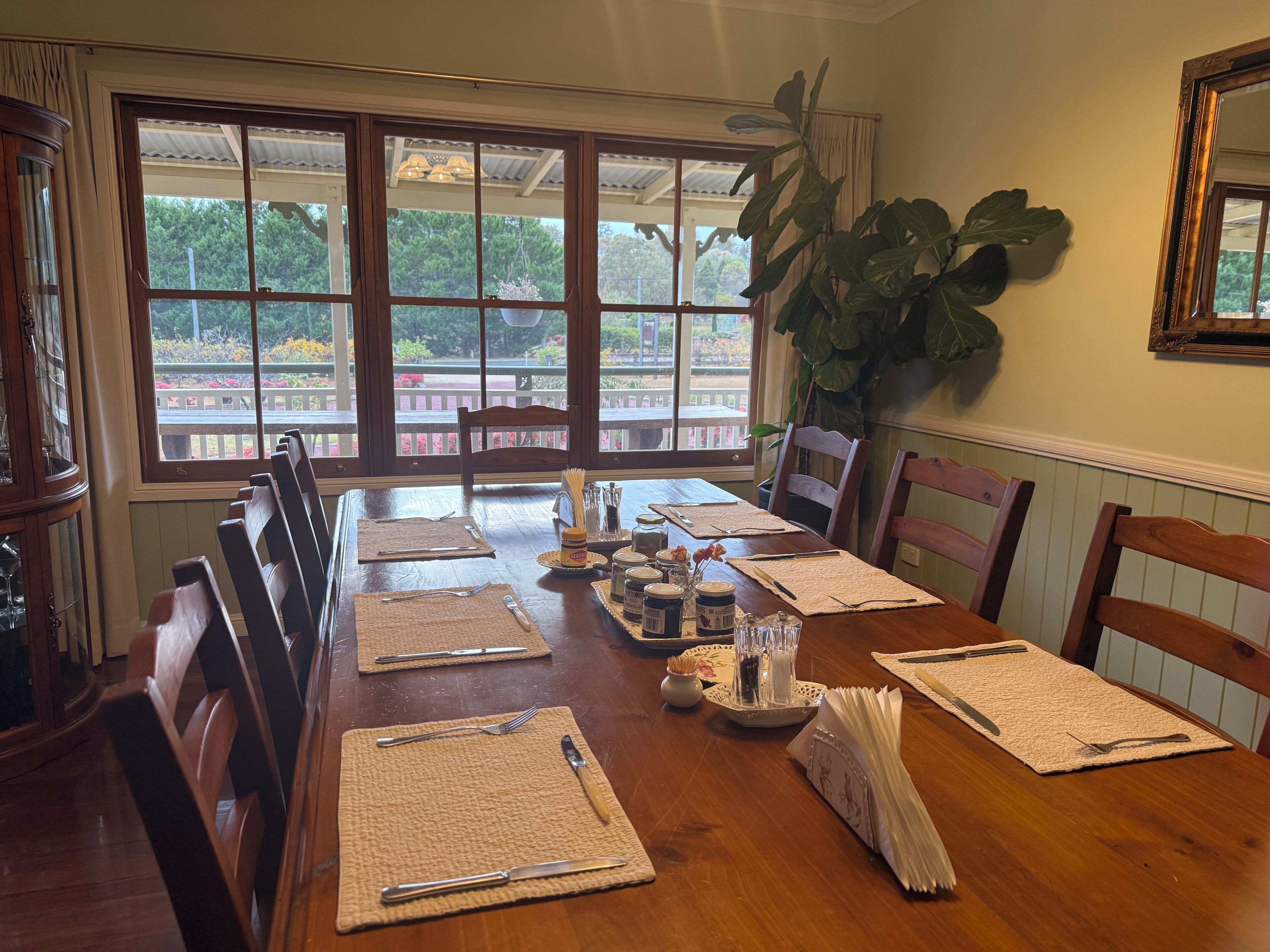 A dining room with a wooden table set for six, large windows, and a view of a garden.