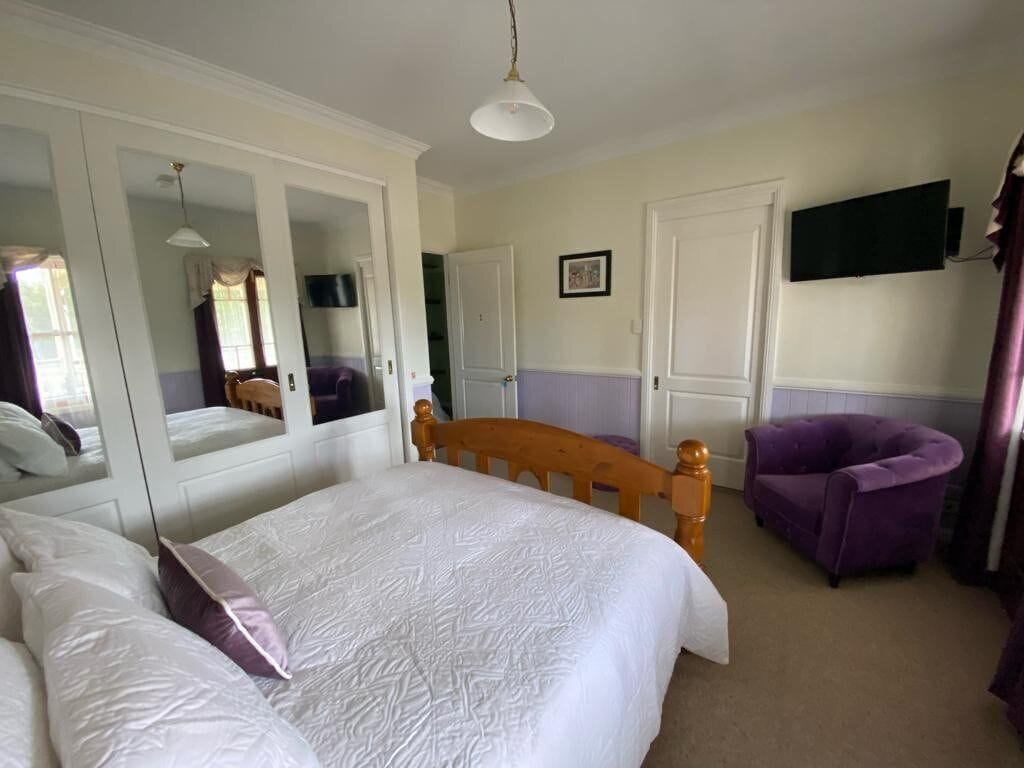 A hotel room with a bed, a wooden headboard, a purple armchair, a television mounted on the wall, and a mirror reflecting part of the room.