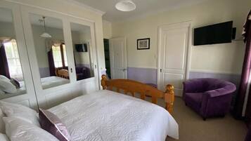 A hotel room with a bed, a wooden headboard, a purple armchair, a television mounted on the wall, and a mirror reflecting part of the room.