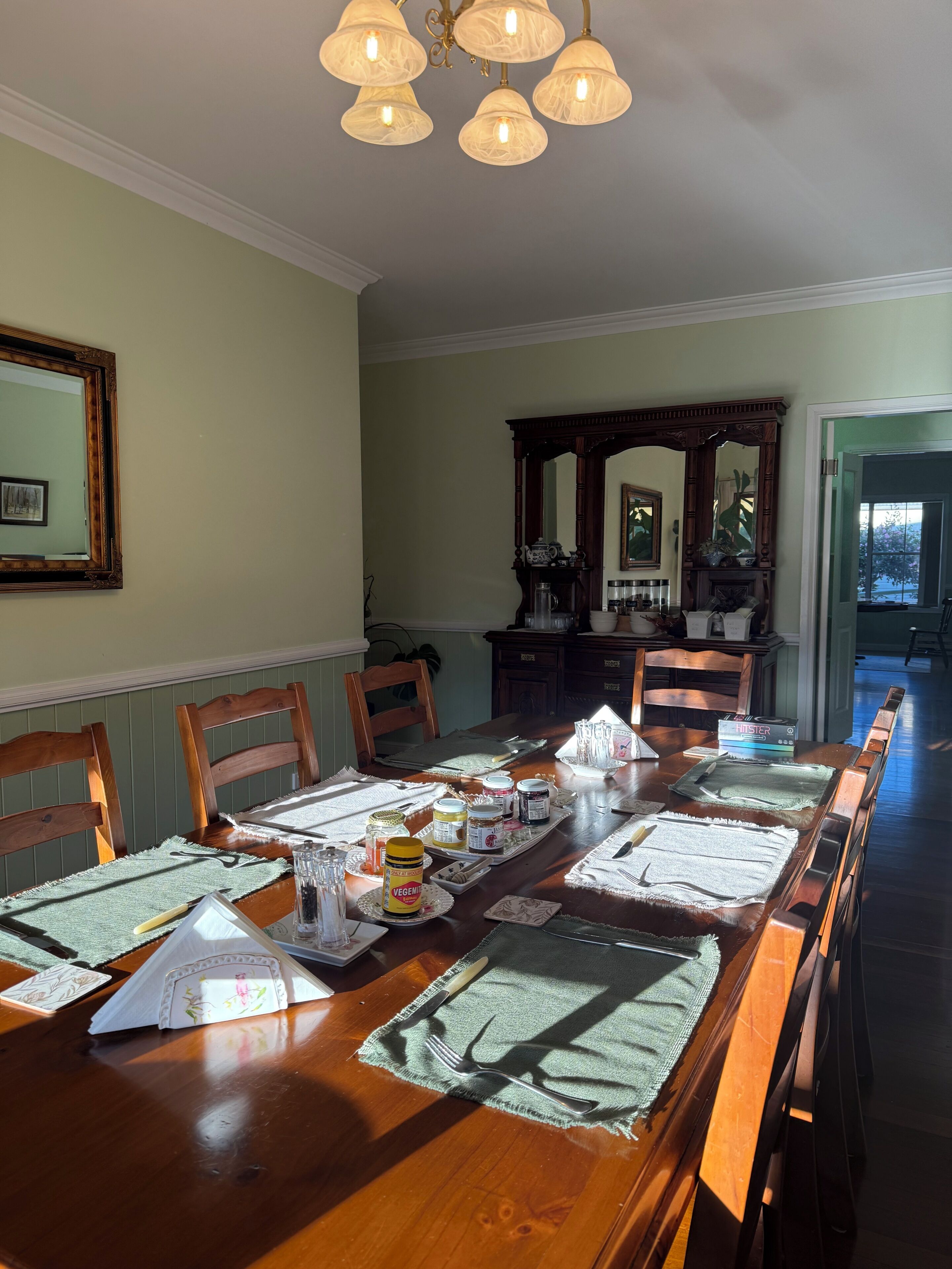Une salle à manger avec une table en bois mise pour le repas, entourée de chaises, un lustre au-dessus et un miroir au mur.
