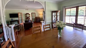 Un comedor con una mesa de madera, sillas y un jarrón de flores. Se observa un televisor al fondo y un piano a la izquierda.