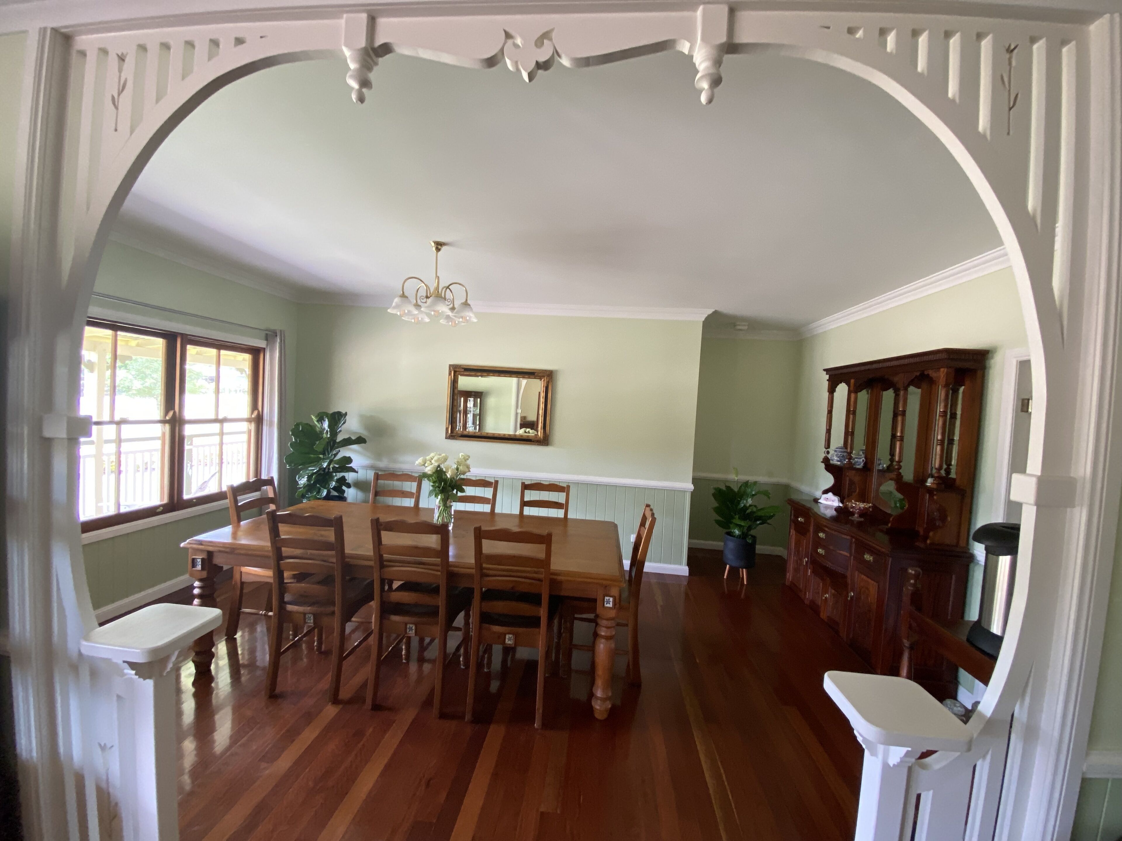 A dining room with a large wooden table, chairs, a chandelier, and a mirror on the wall.