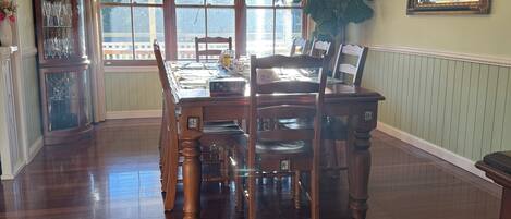 A dining room with a wooden table, chairs, a chandelier, a large window, and a mirror on the wall.