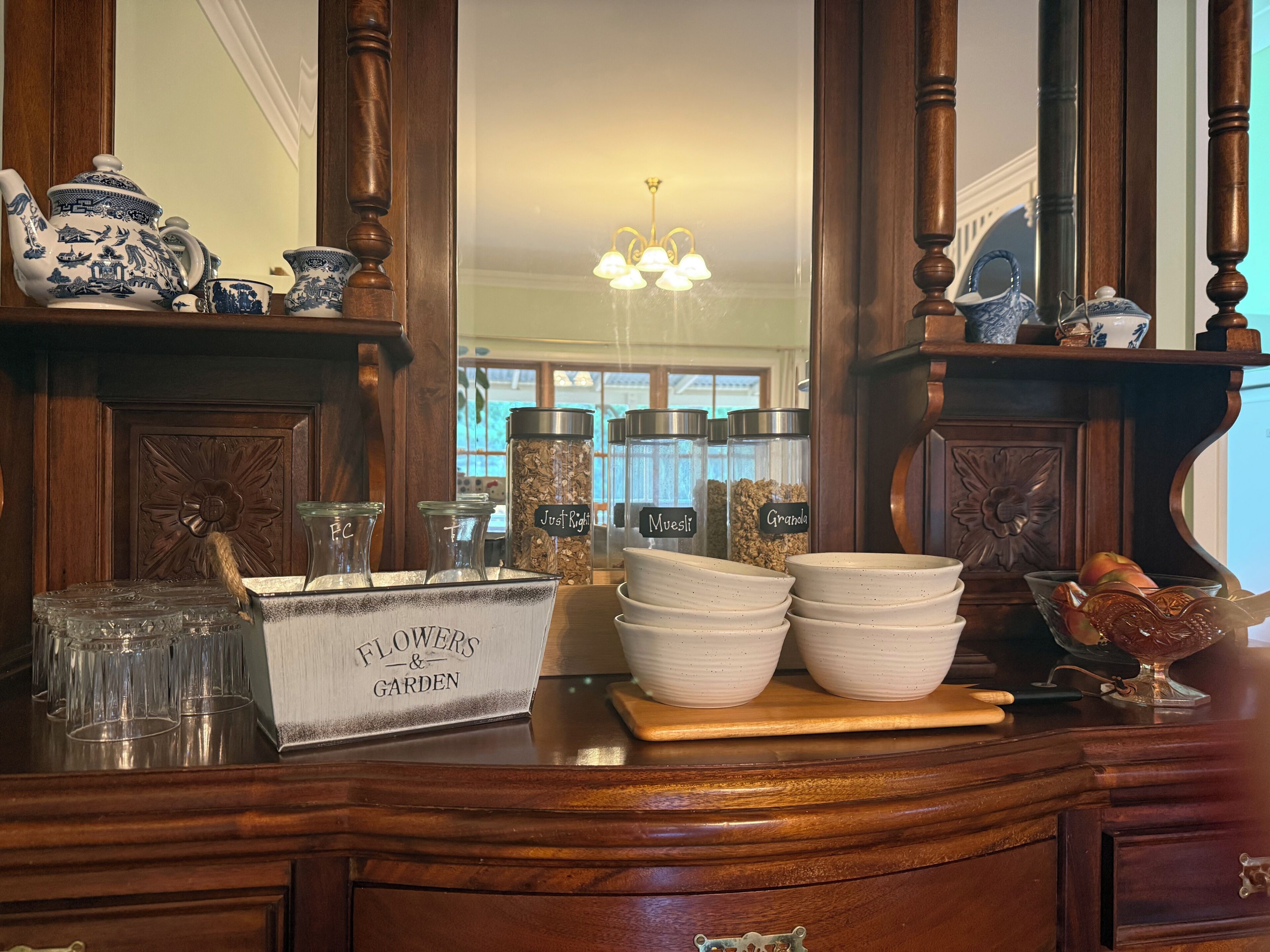 A wooden buffet table with a china set, glass jars labeled with food items, and a sign reading 'FLOWERS & GARDEN'.