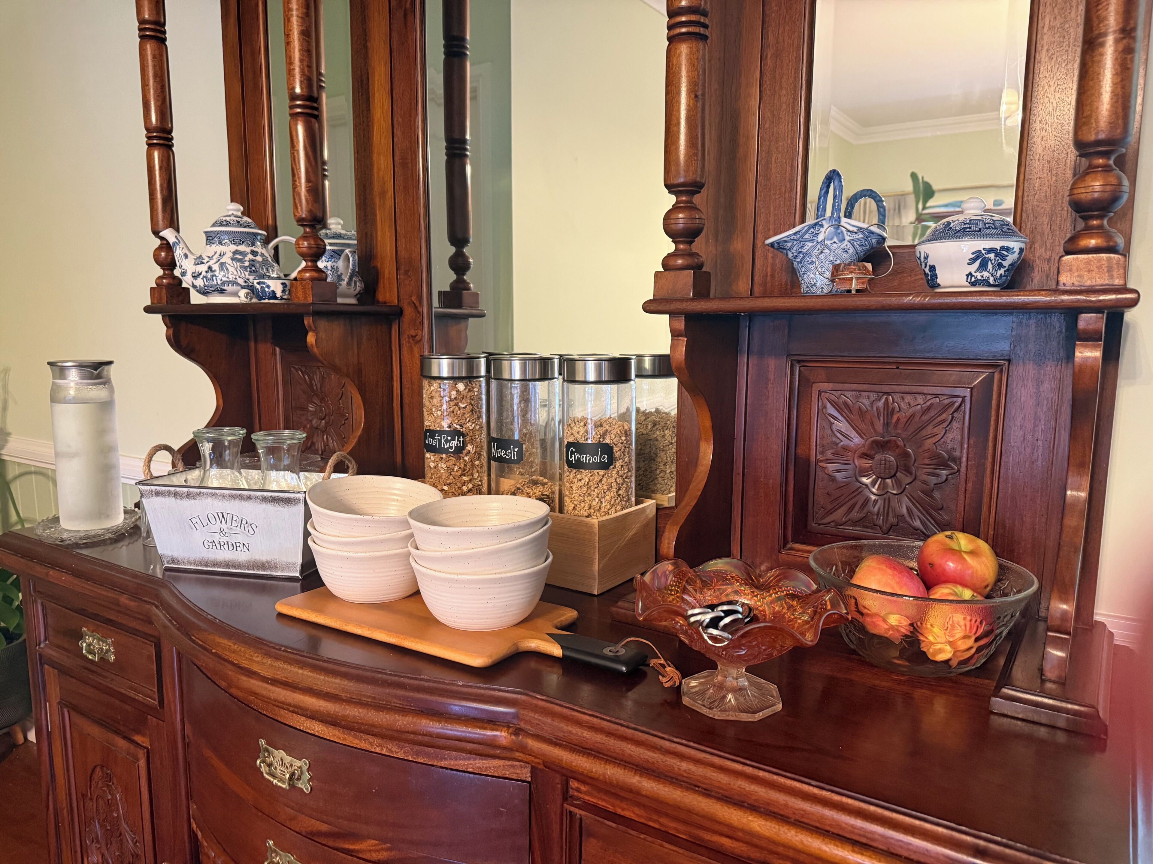 Uma mesa de buffet de madeira com um arranjo floral, uma saladeira e recipientes identificados com granola e outros itens.