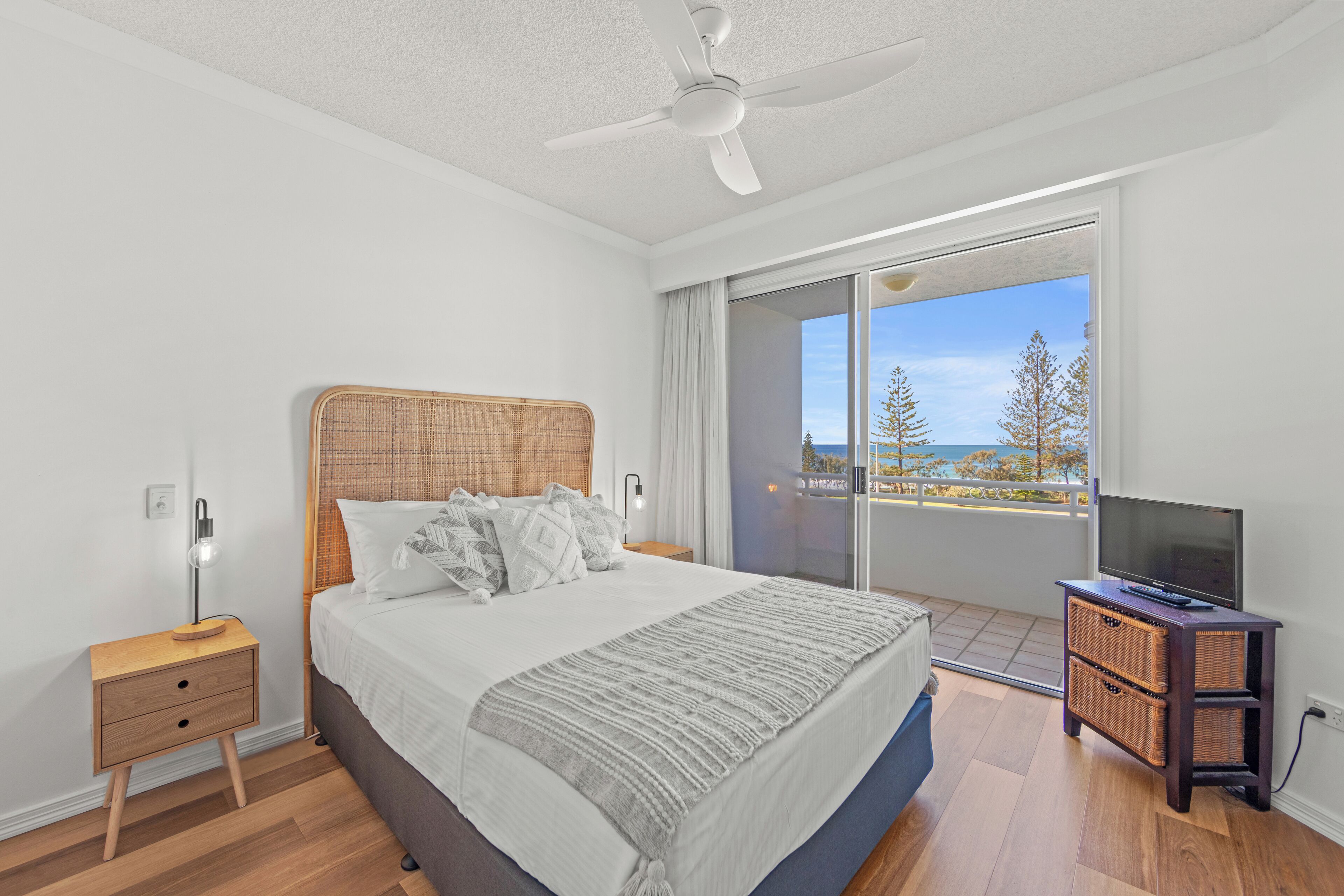 A bedroom with a bed, a nightstand, a television on a stand, and a balcony with a view of trees and a building.