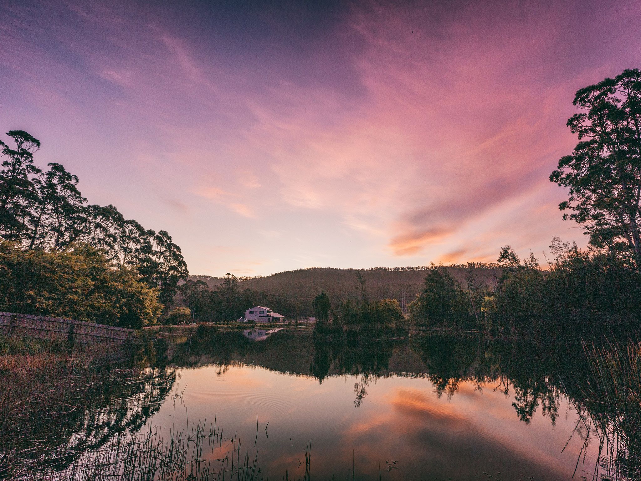 Une vue paisible sur un lac, avec une maison qui se reflète dans l’eau, entourée d’arbres et de collines.