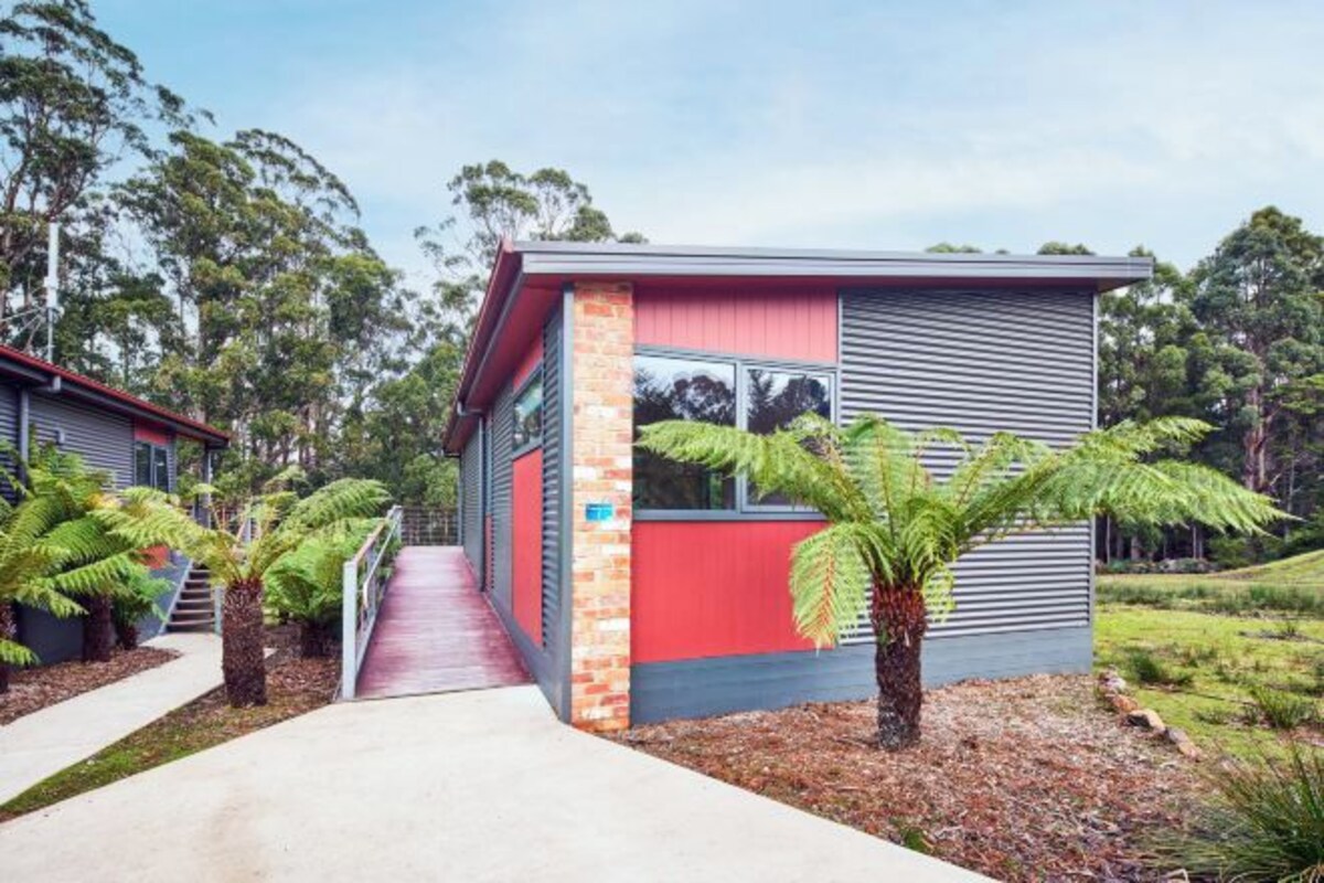 Un bâtiment moderne à la façade rouge et grise, avec un chemin en béton menant à l’entrée, le tout entouré de verdure.