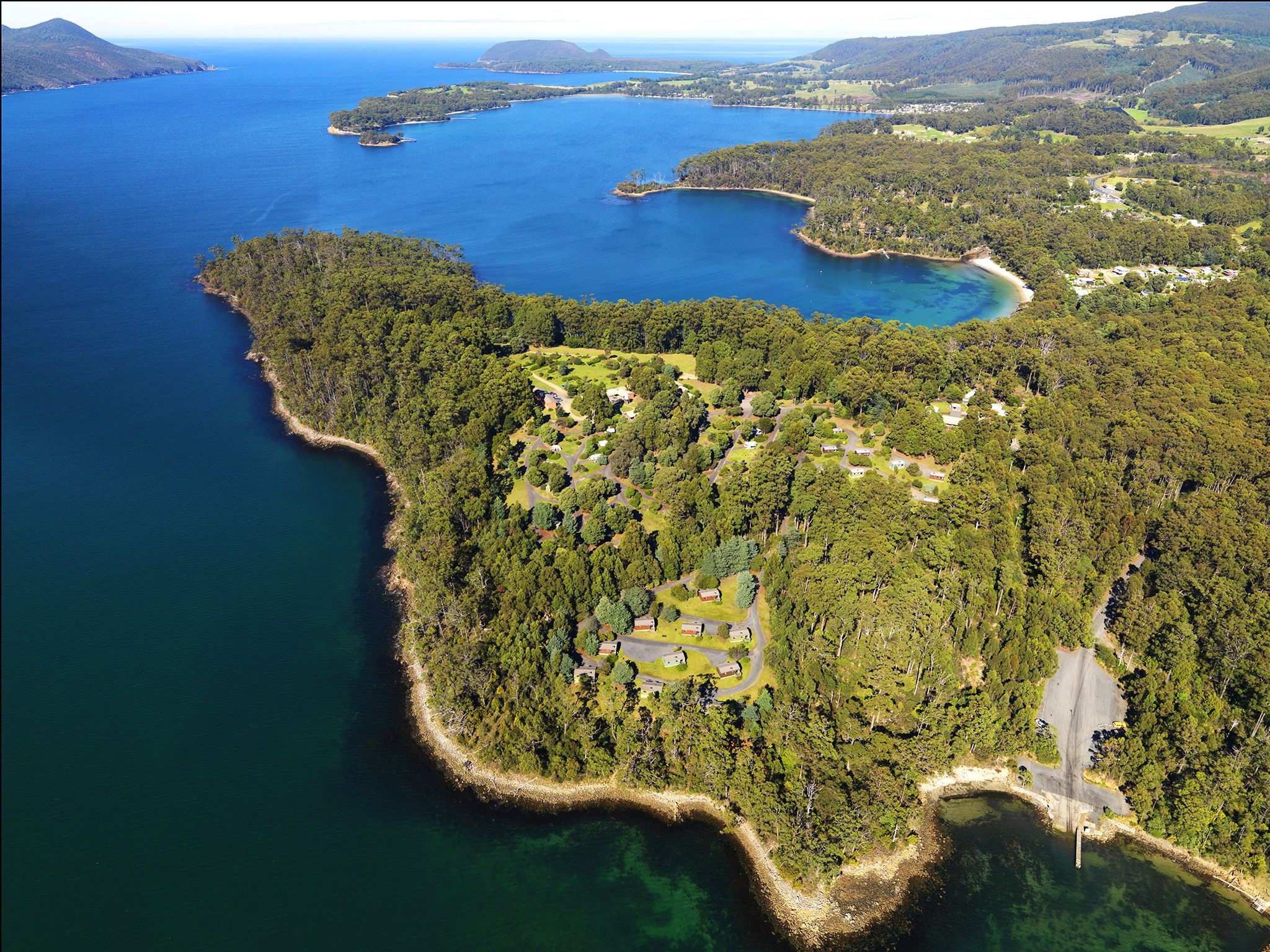 Eine Vogelperspektive zeigt eine Küstenregion mit dichtem Wald, klarem, blauem Wasser und einer kleinen Siedlung mit sichtbaren Straßen und Gebäuden.
