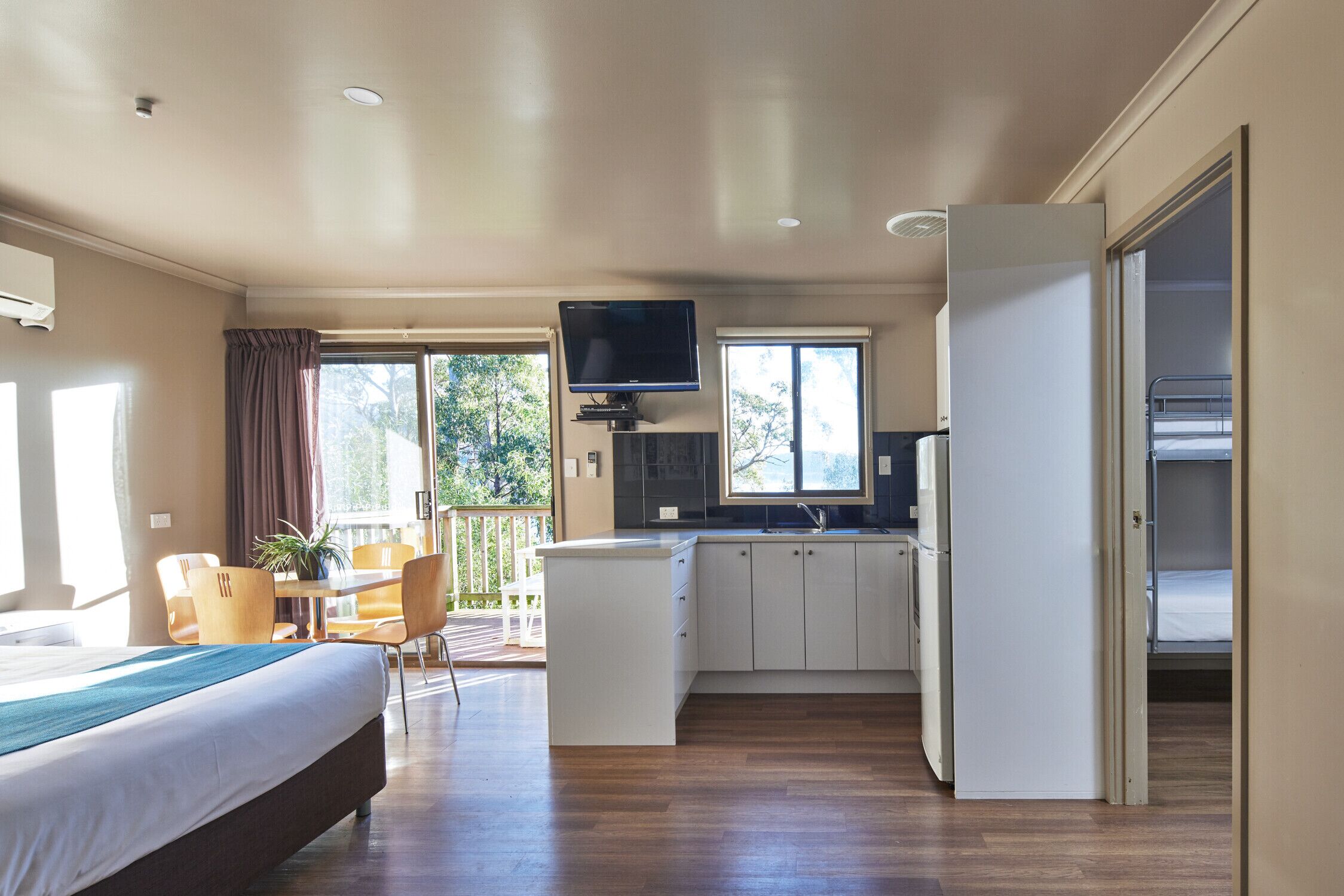 A hotel room with a bed, a dining area with chairs, a kitchen with cabinets, a TV, and a balcony with a view of trees.