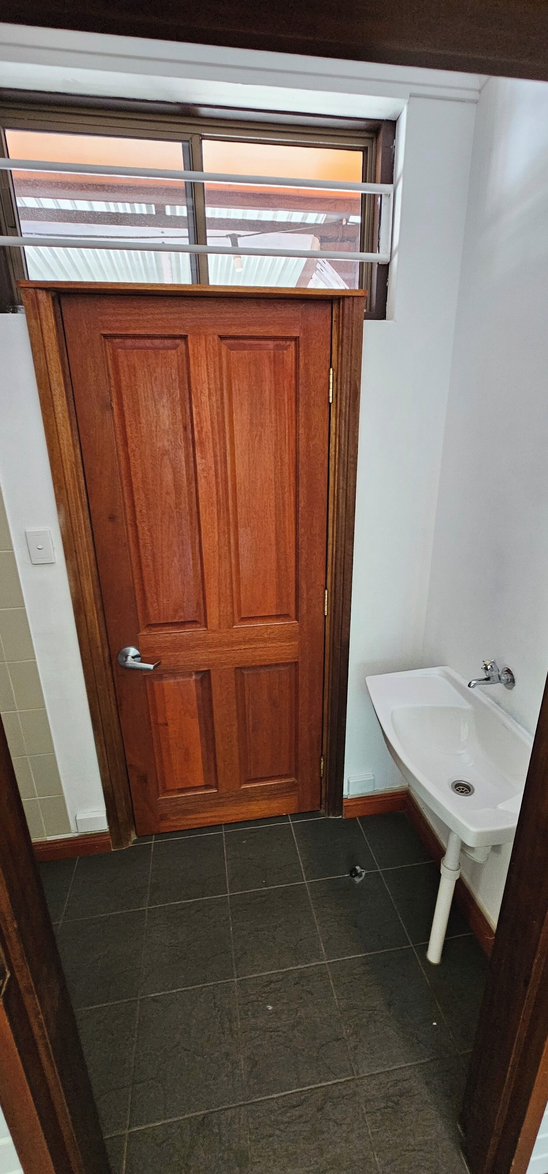 Un baño con puerta de madera, lavamanos blanco y ventana sobre la puerta.